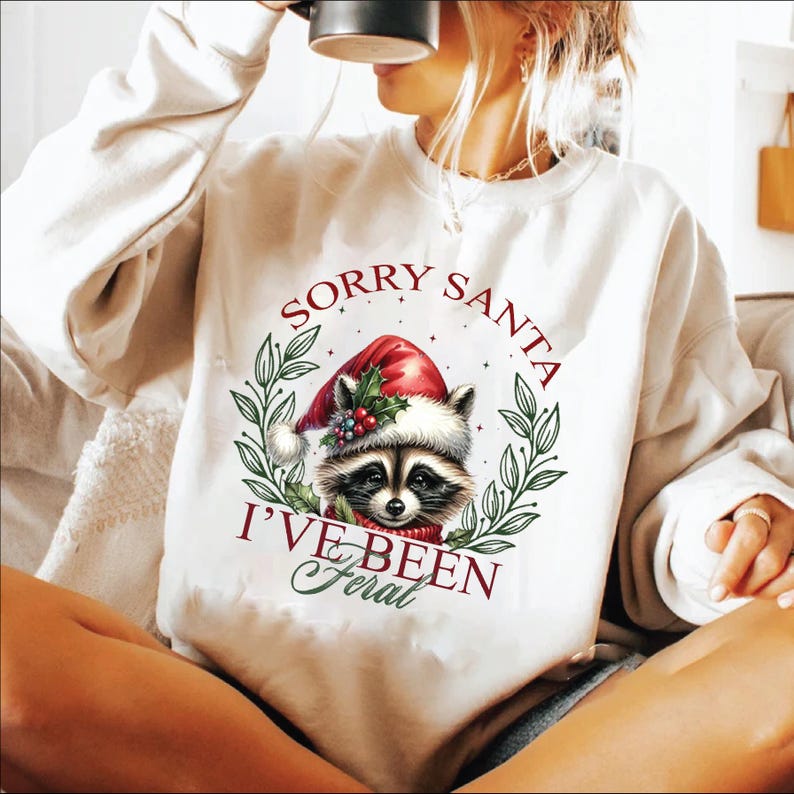 May include: White sweatshirt with a Christmas-themed raccoon wearing a Santa hat and the text "Sorry Santa I've Been Feral".