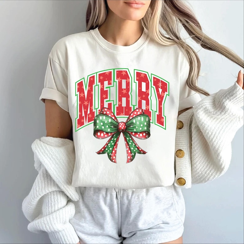 May include: White t-shirt with a red and green "Merry" graphic with a red bow with white polka dots and green leaves.