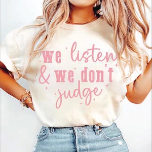 May include: A white t-shirt with a pink graphic that says "We listen & We don't judge".