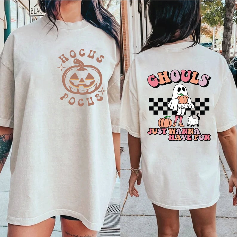 Op de afbeelding: Twee witte T-shirts met Halloween-thema's. Het eerste shirt heeft een pompoen met de tekst "Hocus Pocus". Het tweede shirt heeft een spook dat een pompoen vasthoudt met de tekst "Ghouls Just Wanna Have Fun".