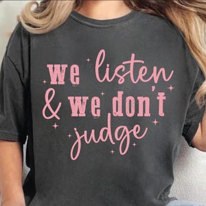 May include: A dark gray t-shirt with the text "We listen & we don't judge" printed in pink.