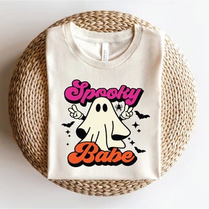 Op de afbeelding: Een wit T-shirt met een retro-grafische afbeelding van een geest met de tekst "Spooky Babe" in roze, oranje en zwart.