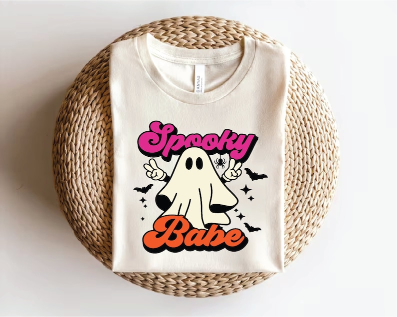 Op de afbeelding: Een wit T-shirt met een retro-grafische afbeelding van een geest met de tekst "Spooky Babe" in roze, oranje en zwart.