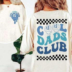 May include: White t-shirt with a blue and white graphic that says "Cool Dads Club" on the front and back. The graphic on the back is larger and has a checkered pattern at the top and bottom.