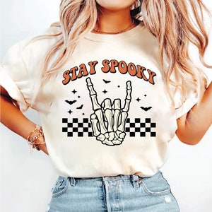 Op de afbeelding: Wit T-shirt met een skelet hand die een rock-and-roll gebaar maakt. Het shirt heeft de tekst "STAY SPOOKY" in oranje letters met een geruit patroon onder de hand.