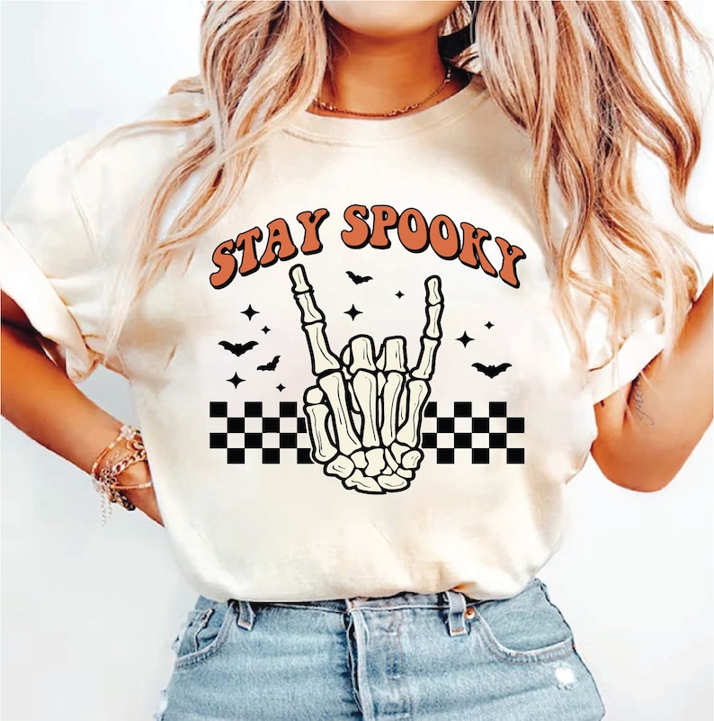 Op de afbeelding: Wit T-shirt met een skelet hand die een rock-and-roll gebaar maakt. Het shirt heeft de tekst "STAY SPOOKY" in oranje letters met een geruit patroon onder de hand.