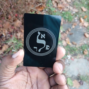 May include: A black rectangular card with a white circular design featuring Hebrew letters. The card is held in a hand against a blurred background of fallen leaves and greenery. The design is centered on the card.