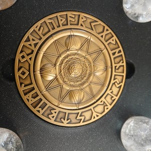 Könnte beinhalten: Ein kreisförmiges, bronzefarbenes Medaillon mit einem komplexen geometrischen Sternmuster in der Mitte, umgeben von einer Umrandung aus eingravierten Runenzeichen. Das Medaillon liegt auf einer schwarzen, strukturierten Oberfläche mit vier klaren Kugeln.