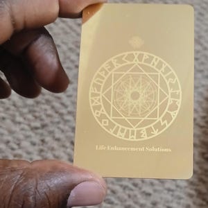 Pode incluir: Um cartão retangular dourado com um design circular com runas e formas geométricas, e o texto "Life Enhancement Solutions". O cartão é segurado por uma pessoa.