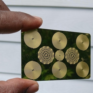 May include: A rectangular, transparent card with a green tint, featuring gold spiral and geometric designs. The card is held between fingers, showcasing the intricate patterns. The background is blurred, suggesting an outdoor setting.