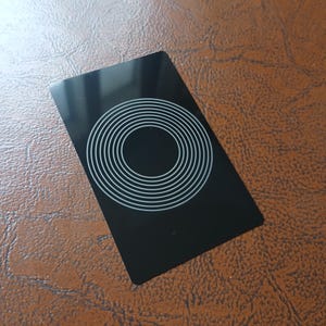 May include: A black rectangular card with a concentric circle design in white. The card is glossy and appears to be made of plastic or a similar material. The background is a textured brown surface.