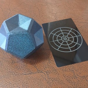 May include: A dark blue, geometric, 12-sided die with a black and white target card.