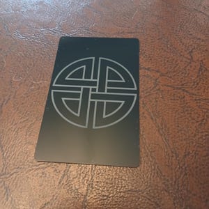 Puede incluir: Una tarjeta rectangular negra con un diseño de nudo celta grabado en un color más claro. La tarjeta está ligeramente inclinada, mostrando el intrincado patrón de nudo. El fondo es una superficie marrón texturizada.