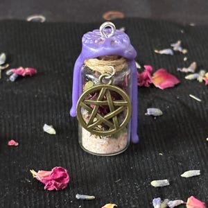 May include: A small glass vial filled with dried herbs and light-colored sand, sealed with a purple wax top featuring a metal loop. A bronze-toned pentacle charm hangs from a twine cord wrapped around the neck of the bottle.