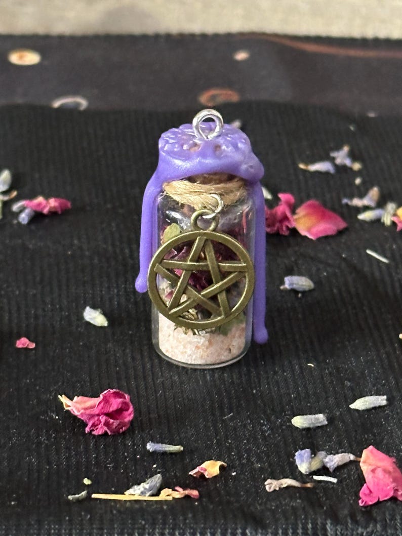 May include: A small glass vial filled with dried herbs and light-colored sand, sealed with a purple wax top featuring a metal loop. A bronze-toned pentacle charm hangs from a twine cord wrapped around the neck of the bottle.
