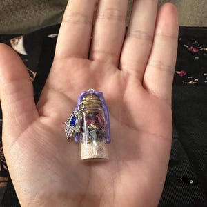 May include: A small clear glass vial filled with layers of sand, dried flower petals, and small stones, sealed with a cork and purple wax. A silver-toned hamsa charm with a blue stone inlay is attached to the top of the glass bottle.