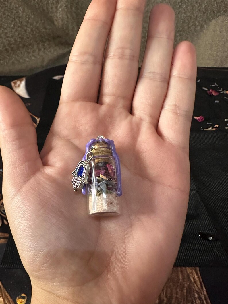 May include: A small clear glass vial filled with layers of sand, dried flower petals, and small stones, sealed with a cork and purple wax. A silver-toned hamsa charm with a blue stone inlay is attached to the top of the glass bottle.