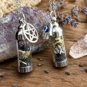 May include: Two silver-toned pendant necklaces featuring clear glass vials filled with dried herbs, small dark stones, and lavender sprigs. One vial has a silver pentagram charm, and the other has a silver hamsa hand charm with a blue evil eye bead.