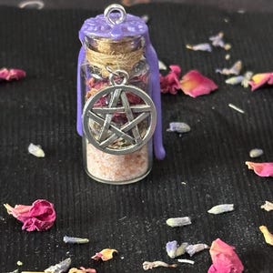 May include: A small clear glass vial filled with pink salt and dried herbs, sealed with a cork and purple wax. A silver-toned metal pentacle charm hangs from the neck of the bottle, which is wrapped in twine and topped with a metal loop.