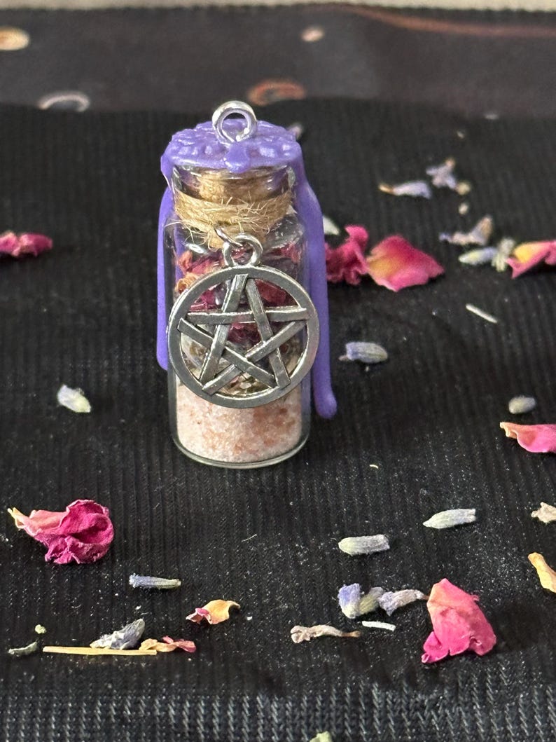May include: A small clear glass vial filled with pink salt and dried herbs, sealed with a cork and purple wax. A silver-toned metal pentacle charm hangs from the neck of the bottle, which is wrapped in twine and topped with a metal loop.