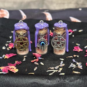 May include: Three small glass vials filled with pink salt and dried flower petals, each sealed with purple wax and a silver jump ring. The vials feature metal charms: a gold pentacle, a silver hamsa hand with a blue eye, and a silver pentacle.