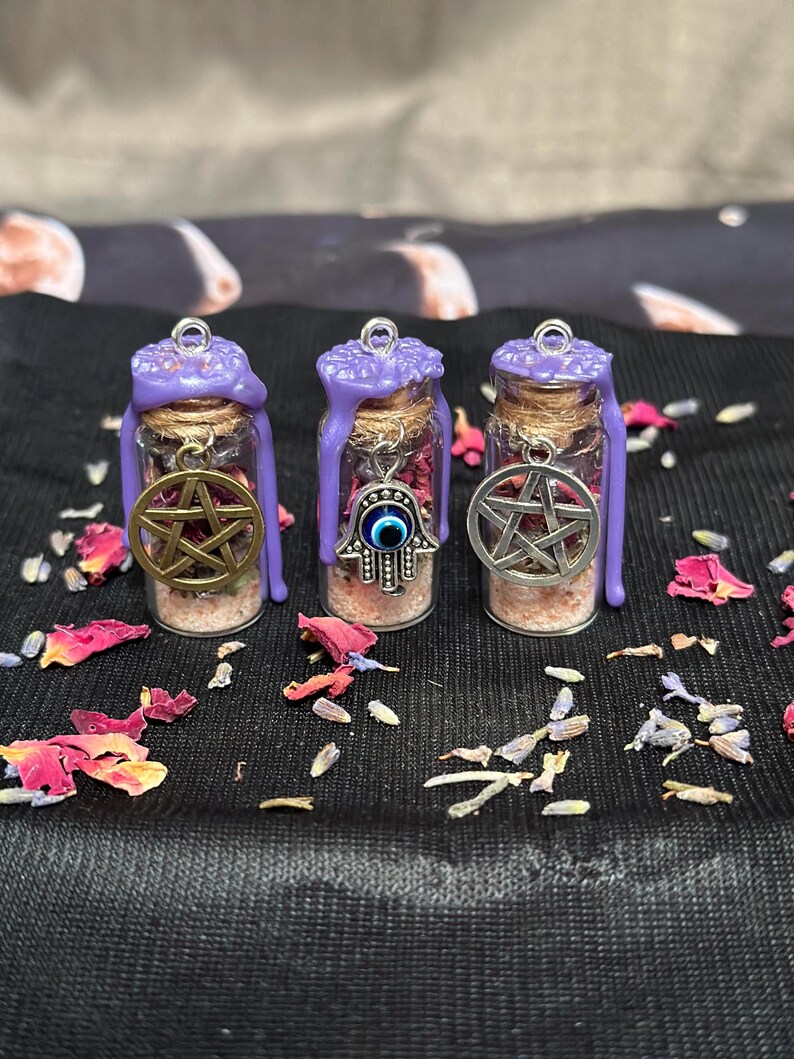 May include: Three small glass vials filled with pink salt and dried flower petals, each sealed with purple wax and a silver jump ring. The vials feature metal charms: a gold pentacle, a silver hamsa hand with a blue eye, and a silver pentacle.