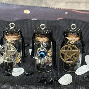 May include: Three small glass bottles with black lids and charms hanging from them. The charms are a pentagram, a hamsa hand with an evil eye, and a pentagram with a gold finish. The bottles are filled with black sand and crystals.