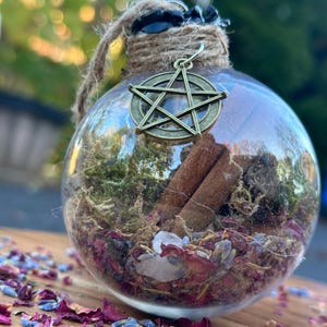 May include: A clear glass orb with a pentagram charm, filled with dried herbs, rose petals, and cinnamon sticks. The orb is tied with twine and sits on a wooden surface with scattered rose petals.