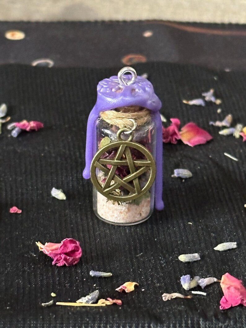 May include: A small glass vial filled with dried herbs and light-colored sand, sealed with a purple wax top featuring a metal loop. A bronze-toned pentacle charm hangs from a twine cord wrapped around the neck of the bottle.