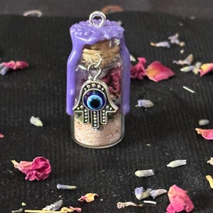 May include: A small glass vial filled with pink sand and dried flower petals, sealed with a cork and topped with purple wax. A silver-toned hamsa charm featuring a blue and white evil eye bead hangs from the neck of the bottle.