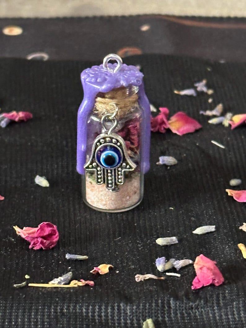 May include: A small glass vial filled with pink sand and dried flower petals, sealed with a cork and topped with purple wax. A silver-toned hamsa charm featuring a blue and white evil eye bead hangs from the neck of the bottle.