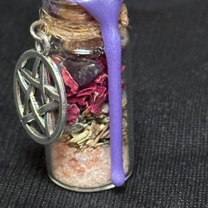 May include: A small glass vial pendant filled with layers of pink salt, dried herbs, red flower petals, and dark stones. The cork stopper is sealed with purple wax that drips down the side. A silver-toned pentacle charm hangs from the neck.