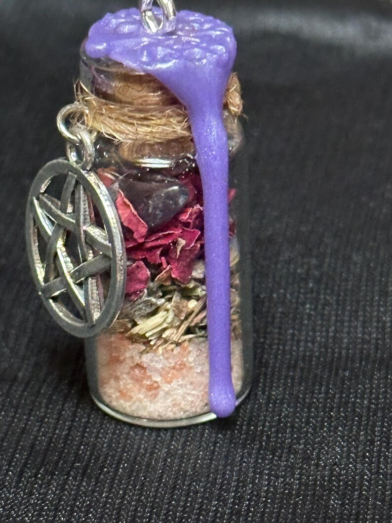 May include: A small glass vial pendant filled with layers of pink salt, dried herbs, red flower petals, and dark stones. The cork stopper is sealed with purple wax that drips down the side. A silver-toned pentacle charm hangs from the neck.