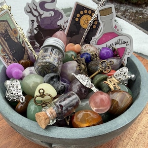 May include: A gray stone bowl filled with assorted polished gemstones, small glass jars containing dried herbs and petals, metal keys, mushroom-shaped charms, and decorative stickers featuring a black cat, a cauldron, and a love potion bottle.