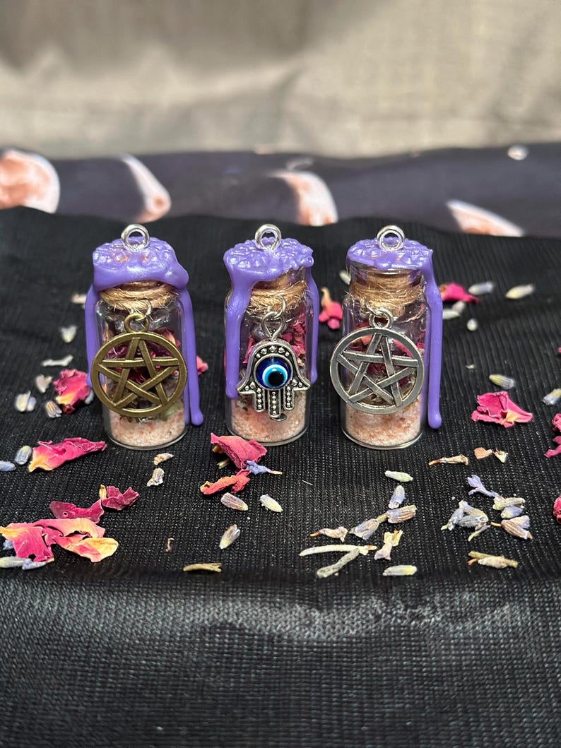 May include: Three small glass bottles with cork stoppers sealed with purple wax. Each bottle contains pink salt and dried flower petals. The bottles feature silver-toned metal charms: two pentacles and one hamsa hand with a blue evil eye bead.