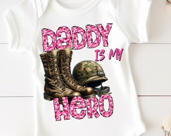 Könnte beinhalten: Weißer Baby-Strampler mit einem pinkfarbenen Tarnmuster, auf dem "Daddy is my Hero" steht. Das Design enthält auch ein Paar braune Militärstiefel und einen grünen Militärhelm.