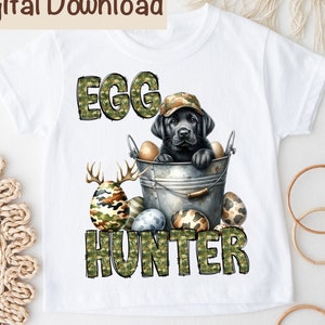 Puede incluir: Camiseta blanca con una ilustración en blanco y negro de un perro con un sombrero de camuflaje y sosteniendo un cubo de huevos de Pascua de camuflaje. El texto "Egg Hunter" está escrito en letras de camuflaje debajo del perro.