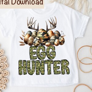 May include: White t-shirt with a camouflage print design featuring a pair of antlers and a pile of Easter eggs. The text "Egg Hunter" is written in a camouflage font below the eggs.