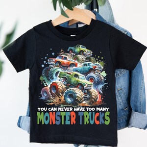 May include: Black t-shirt featuring a colorful graphic of monster trucks in various colors, including green, orange, and blue. The text "YOU CAN NEVER HAVE TOO MANY MONSTER TRUCKS" is printed below the trucks.