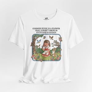 May include: White t-shirt with a colourful illustration inside a blue frame. The image shows a girl with a flower, surrounded by butterflies, birds, a raccoon, and a rabbit. The text reads: "COMMON SENSE IS A FLOWER THAT DOESN'T GROW IN EVERYONE'S GARDEN."