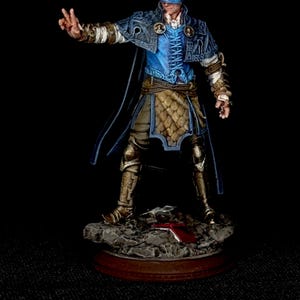 May include: A miniature figurine of a man in fantasy attire. He wears a blue tunic, a gold-colored skirt, and a blue cape. A blue blindfold covers his eyes. The figure is standing on a rocky base with a red object.