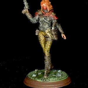 Puede incluir: Figura en miniatura detallada de una guerrera, vestida con un atuendo oscuro y texturizado con detalles rojos en los hombros y una cubierta facial. Sostiene una pistola futurista y está de pie sobre una base con hierba y rocas.