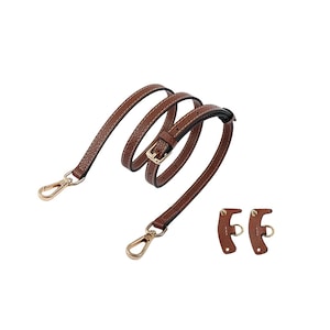 May include: A brown leather purse strap with gold-colored metal clasps and buckle. The strap is stitched with white thread and includes two additional leather pieces with metal rings.