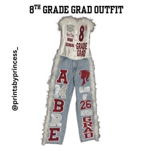 Könnte beinhalten: Ein Outfit für die 8. Klasse. Das Ensemble umfasst ein weißes Oberteil im Korsett-Stil mit rotem Text und eine Jeans aus hellem Denim. Die Jeans zeigen den Namen "AMBRE", eine Abschlusskappe und den Text "CLASS OF 26 8TH GRADE GRAD".