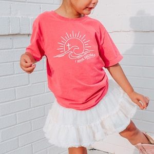 May include: A young girl wearing a pink t-shirt with a white graphic of a sun, wave, and cross. The graphic says "I have sandals". She is wearing a white skirt and pink cowboy boots. She is standing on a brick staircase.