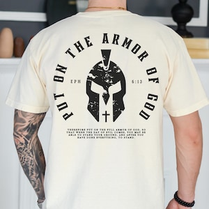 May include: A white t-shirt with a black graphic of a Spartan helmet and the text "Put on the Armor of God" with the scripture reference "Eph 6:13".