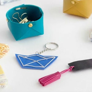 Op de afbeelding: Een assortiment aan knutselbenodigdheden, waaronder polymer clay, een klein blauw leren bakje, een blauwe en witte origami boot sleutelhanger, een roze kwast en een zwart en roze leren band. De afbeelding bevat ook een bord met de tekst "130°C 265°F 30 min."