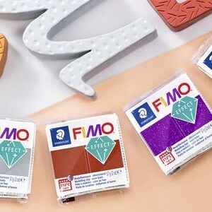 Op de afbeelding: Vier verpakkingen FIMO Effect modelleerklei. De verpakkingen zijn wit met blauwe en groene accenten. De klei kleuren zijn zilver, paars, bruin en goud. Elke verpakking heeft de tekst "FIMO Effect" en "Staedtler" erop gedrukt.