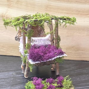 May include: A miniature fairy garden bench made of twigs, moss, and purple and green decorative elements. The bench has a small seat with a purple moss cushion and a green moss roof. The bench is a whimsical decorative item.