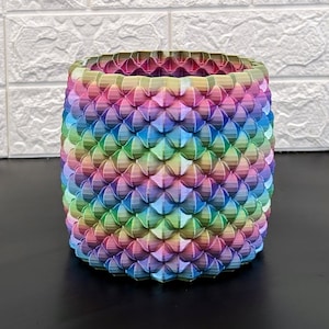 Puede incluir: Un macetero de color arcoíris con un diseño geométrico único. El macetero está hecho de un material texturizado y tiene una forma cilíndrica.